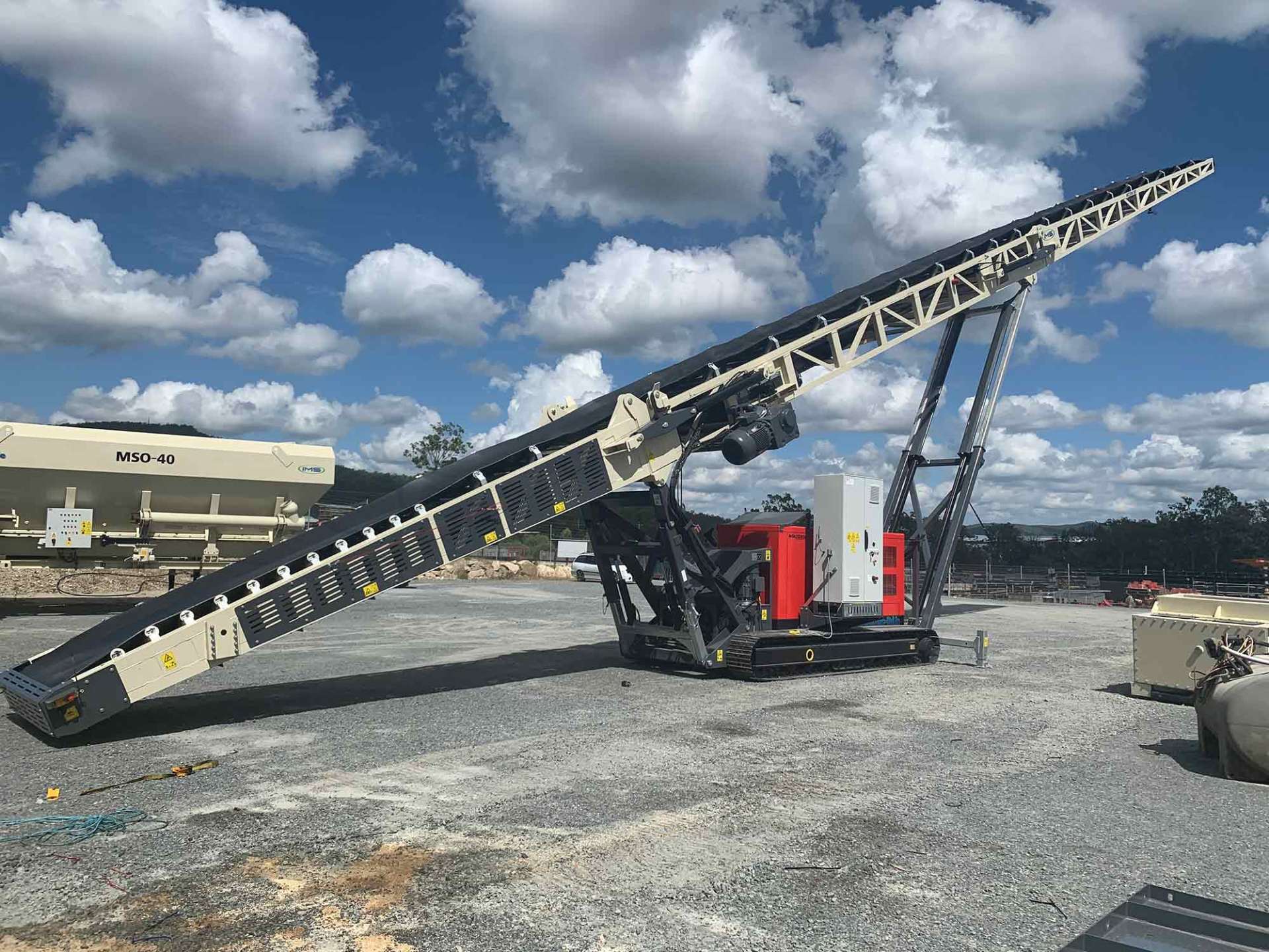 A large mobile conveyor belt, black and white, is angled upwards against a bright blue sky with clouds. It's on a gravel surface.