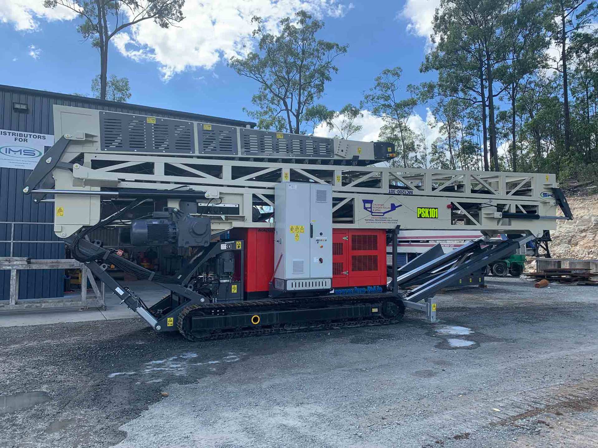 A large, mobile rock crusher on treads with a red and white engine, operating outdoors near a building and trees.