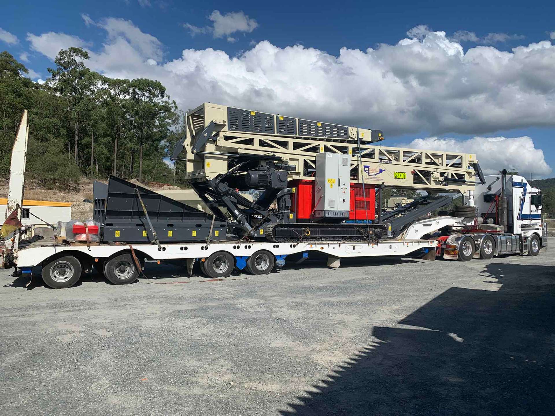 Large industrial machine on a flatbed trailer, likely for crushing rock, parked on gravel under a partly cloudy sky.
