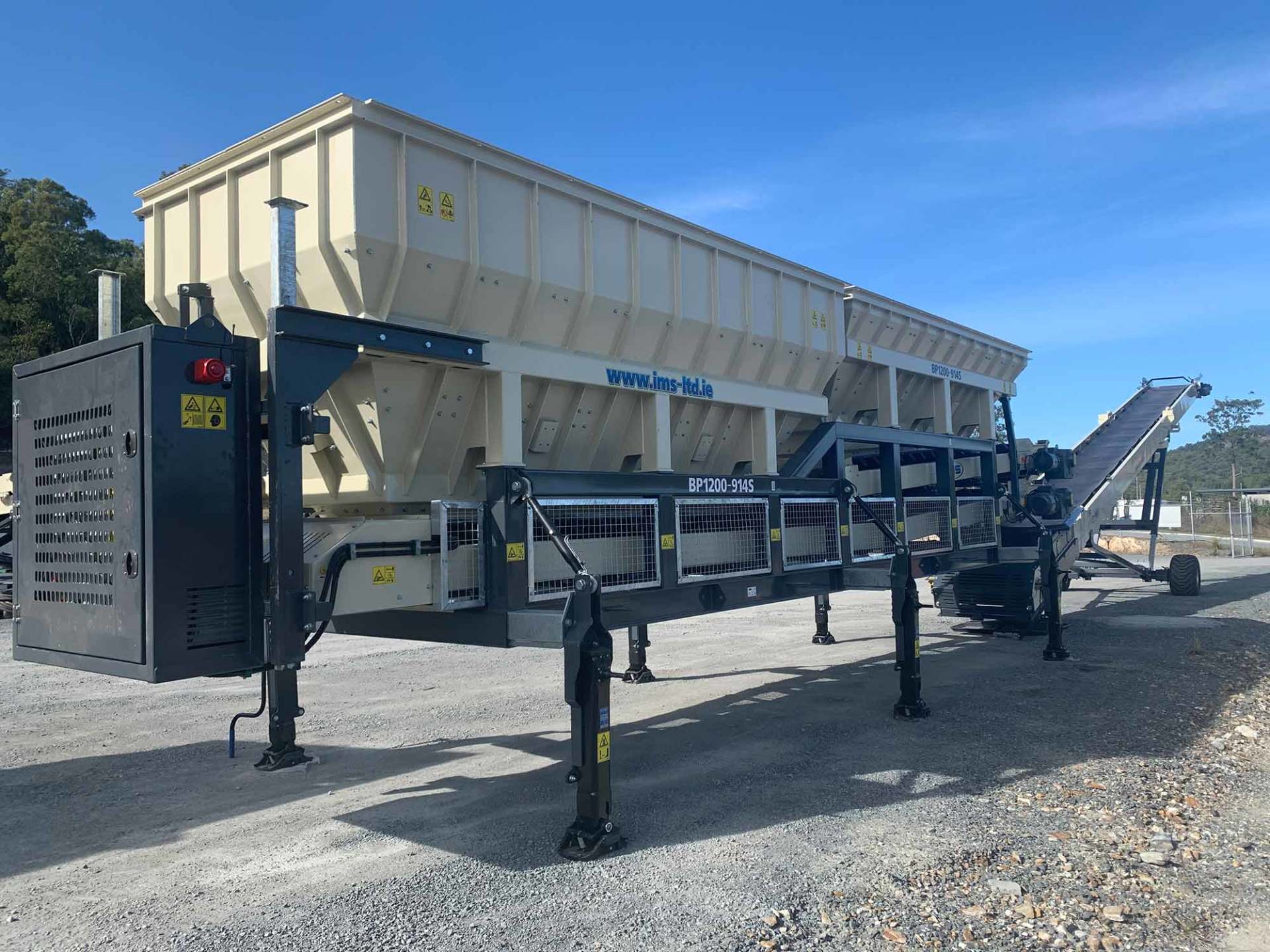Mobile concrete batching plant on a gravel surface. A large beige hopper sits atop a black metal frame with a conveyor belt.