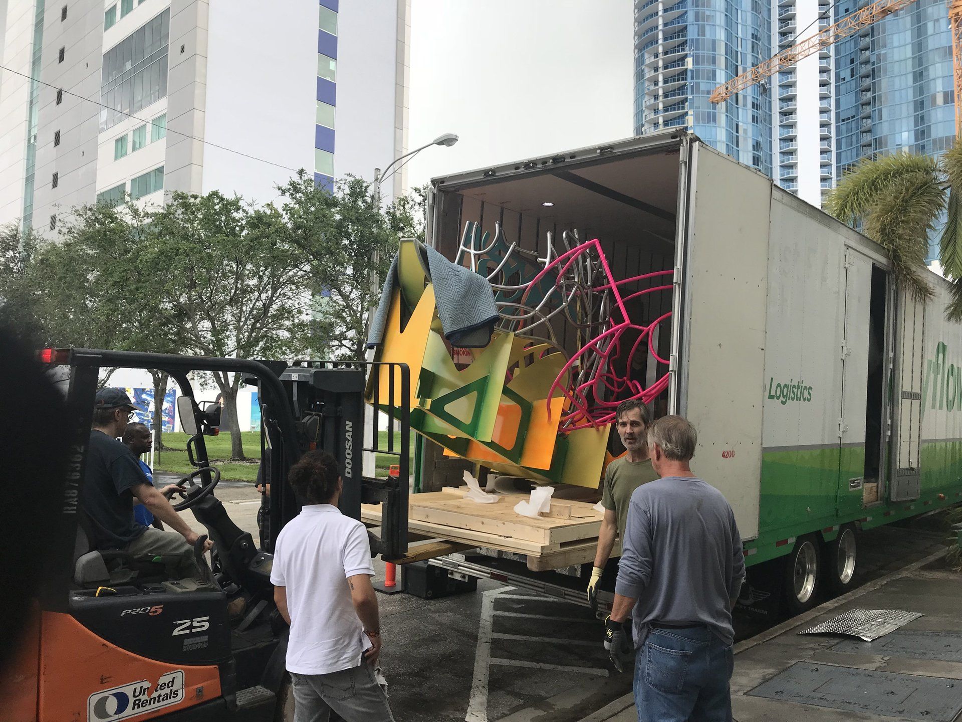 Truck unloading colorful metal frames on a city street, with workers nearby and a forklift assisting