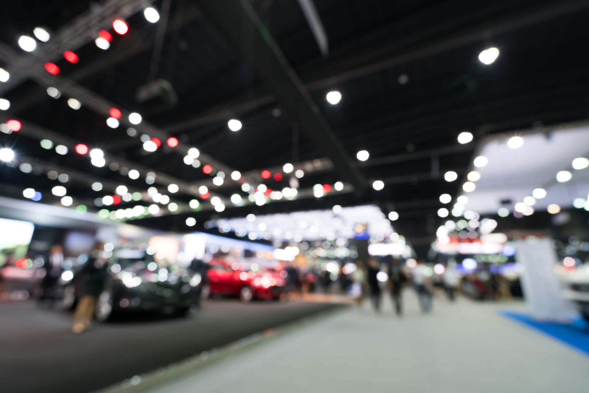 Blurry indoor auto show with cars, bright ceiling lights, and people walking through the exhibition hall