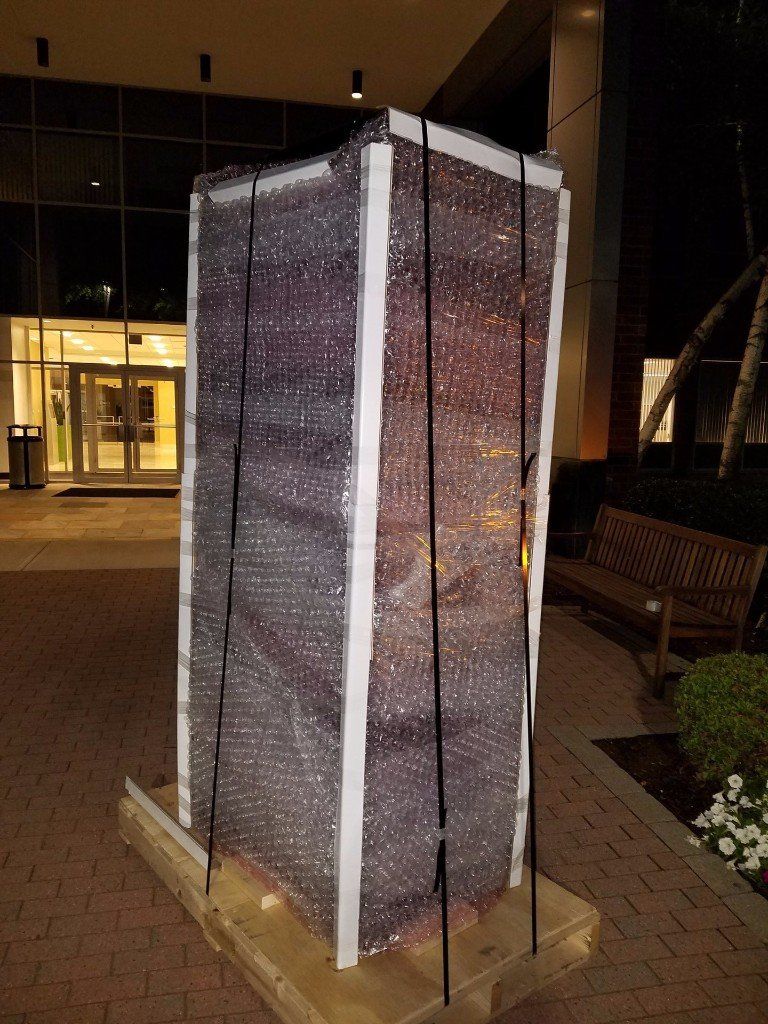 Illuminated fabric-covered rectangular sculpture or booth on a brick walkway at night near a building entrance
