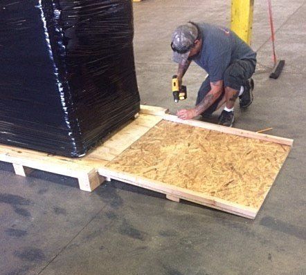 Worker installing plywood ramp beside a large wrapped pallet in a warehouse