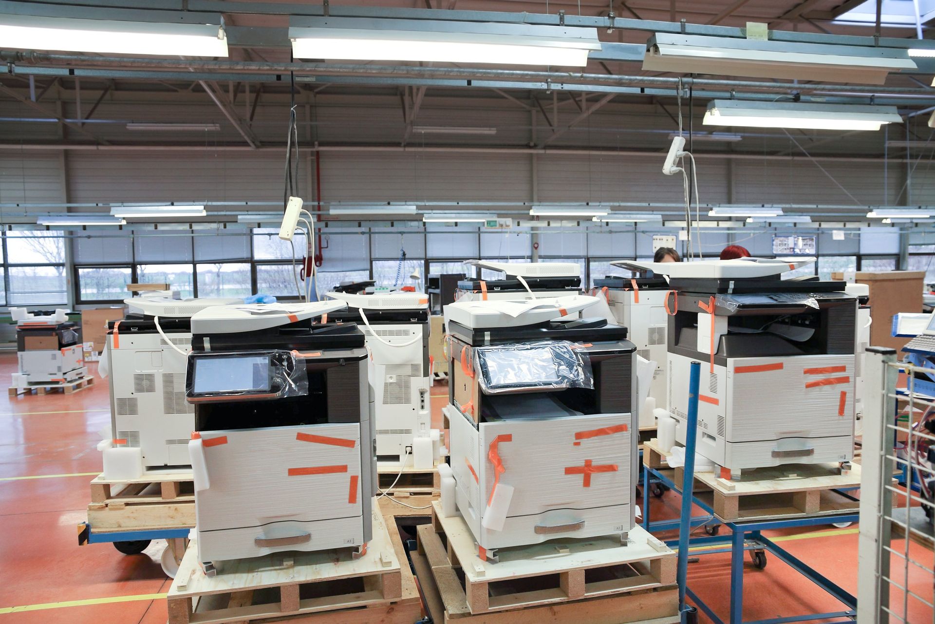Several white office machines wrapped in plastic sit on wooden pallets inside a warehouse.
