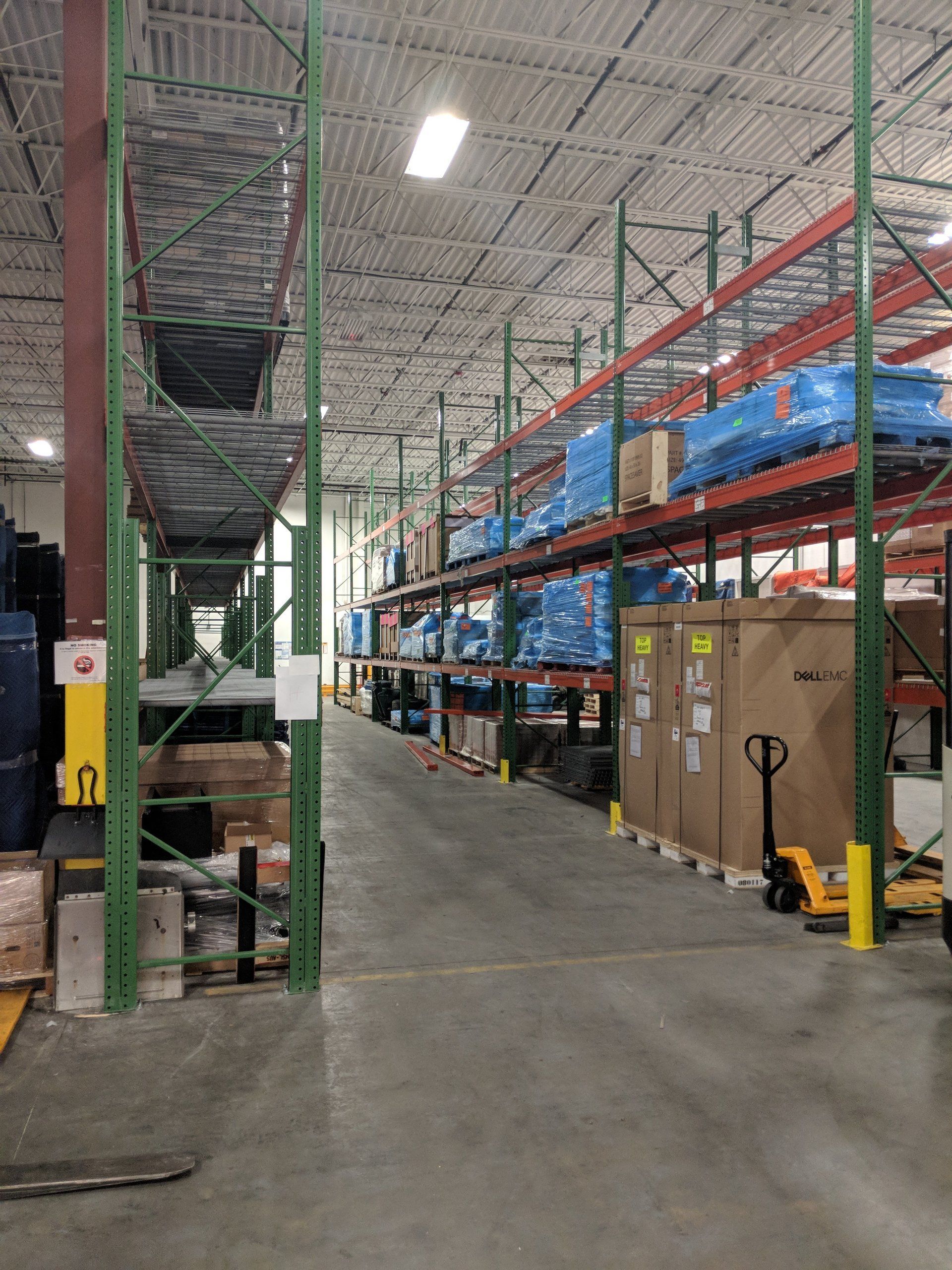 Warehouse aisle with tall racks, blue shrink-wrapped pallets, and a concrete floor under bright overhead lights