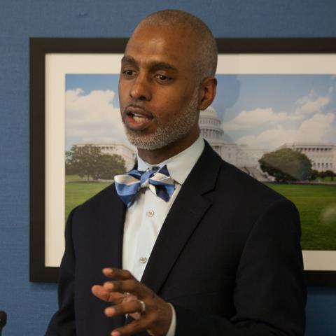 A man in a suit and bow tie stands in front of a picture of the capitol building