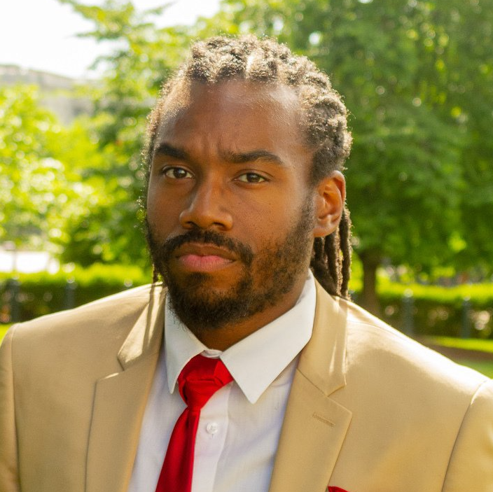 A man with dreadlocks is wearing a tan suit and red tie