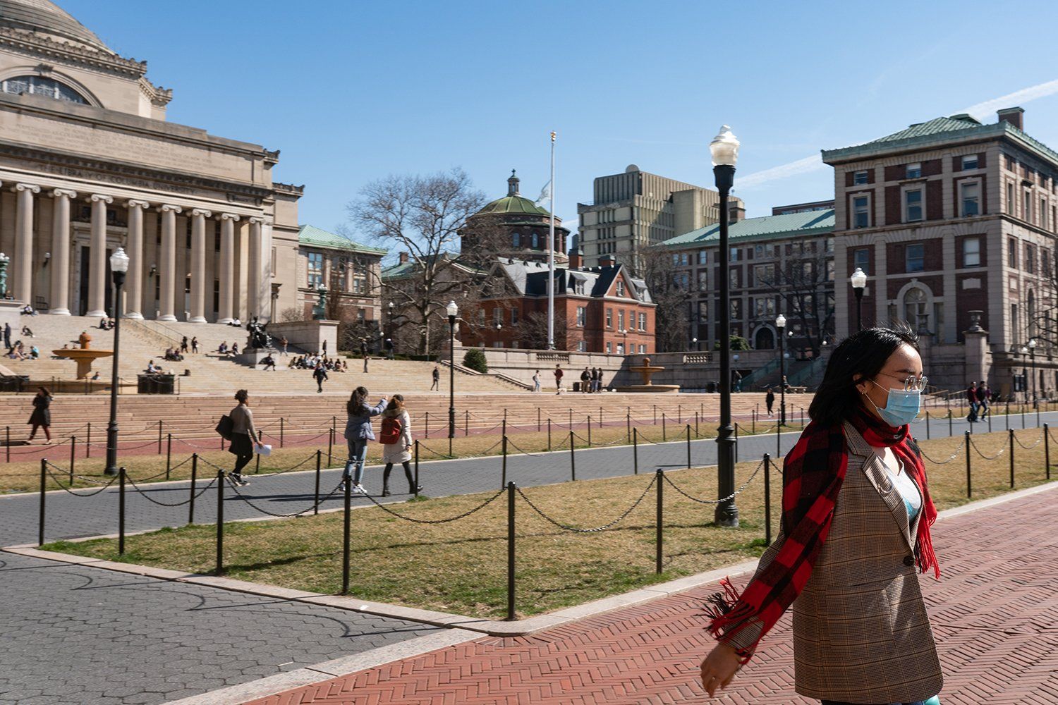 A woman wearing a mask is walking down a sidewalk in front of a large building.