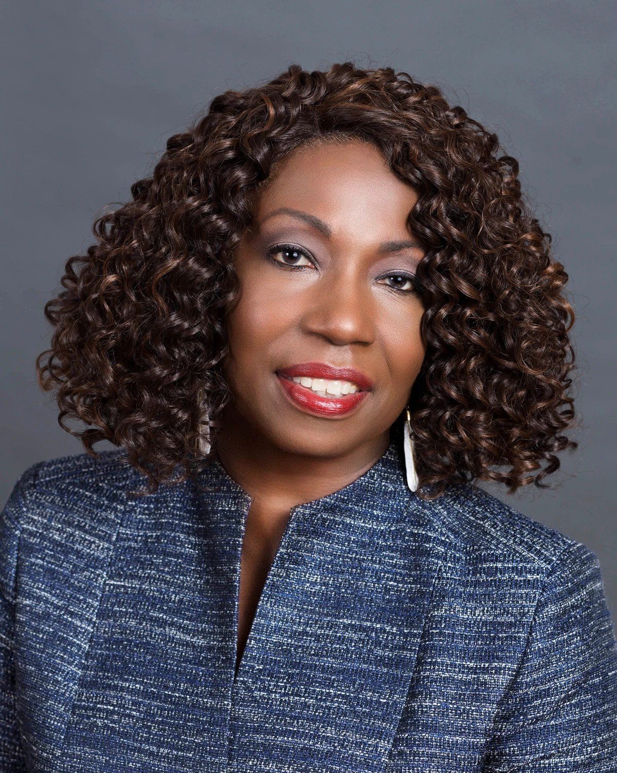 A woman with curly hair is wearing a blue jacket and earrings.
