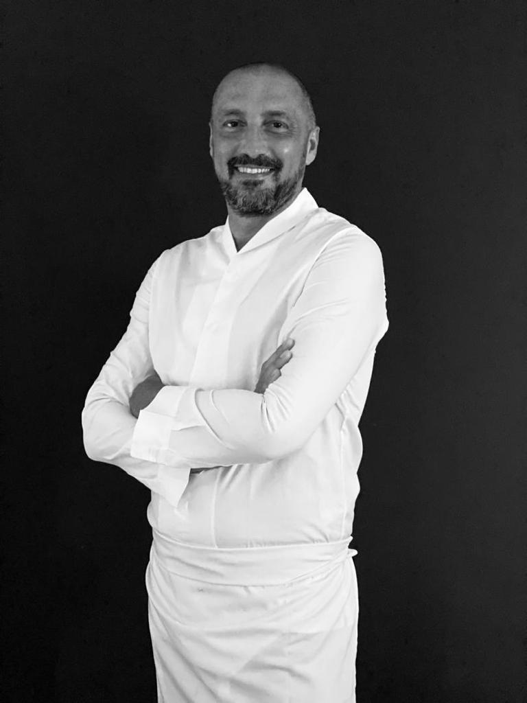 Un homme avec une barbe est debout avec les bras croisés sur une photo en noir et blanc.