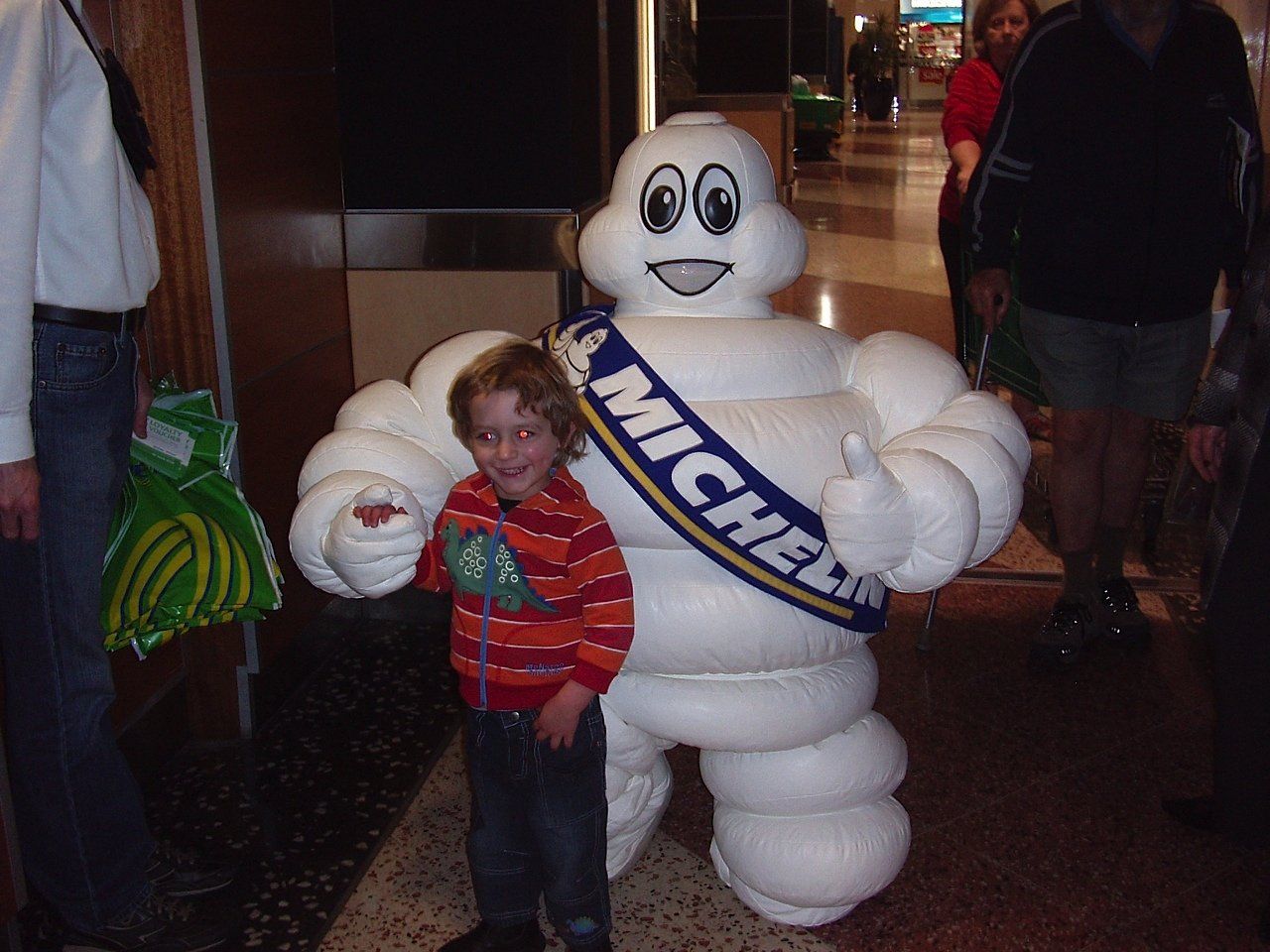 A child smiles next to the Michelin Man mascot, both in an indoor setting. The mascot gives a thumbs-up.
