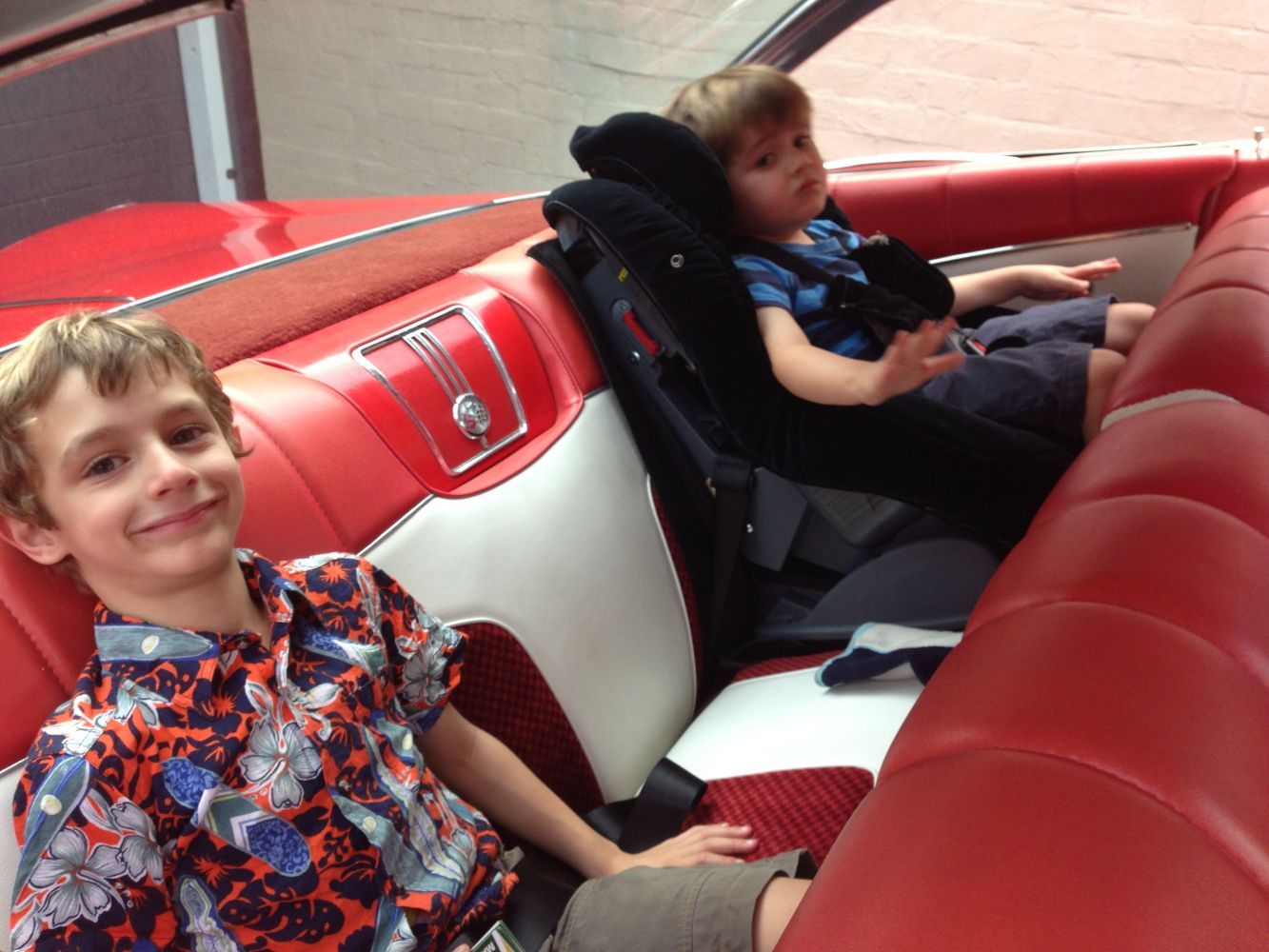 Two children sit in a classic car's red and white interior. One smiles; the other is in a car seat.