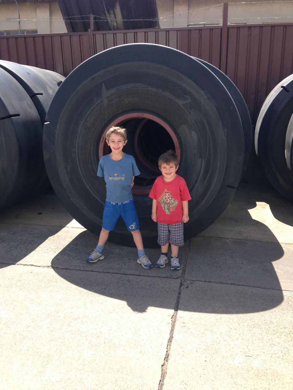 Two children standing inside a massive tire. Outdoors, concrete, sunny.