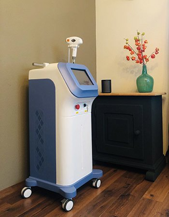 Een wit-blauwe, esthetische lasermachine op wielen staat op een houten vloer naast een donkere kast met een vaas bloemen.