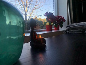 Een zittend Boeddhabeeld met een brandende kaars staat op een donkere tafel bij een raam met rode bloemen en uitzicht op bomen.