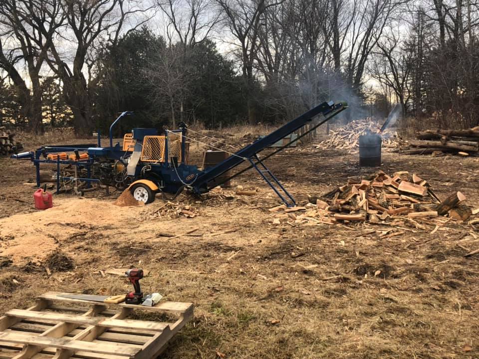 A machine is cutting wood in a field next to a pile of wood.