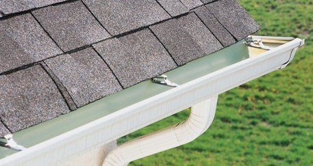White rain gutter attached to a roof edge with shingles and metal mounting clips above a green grassy lawn.