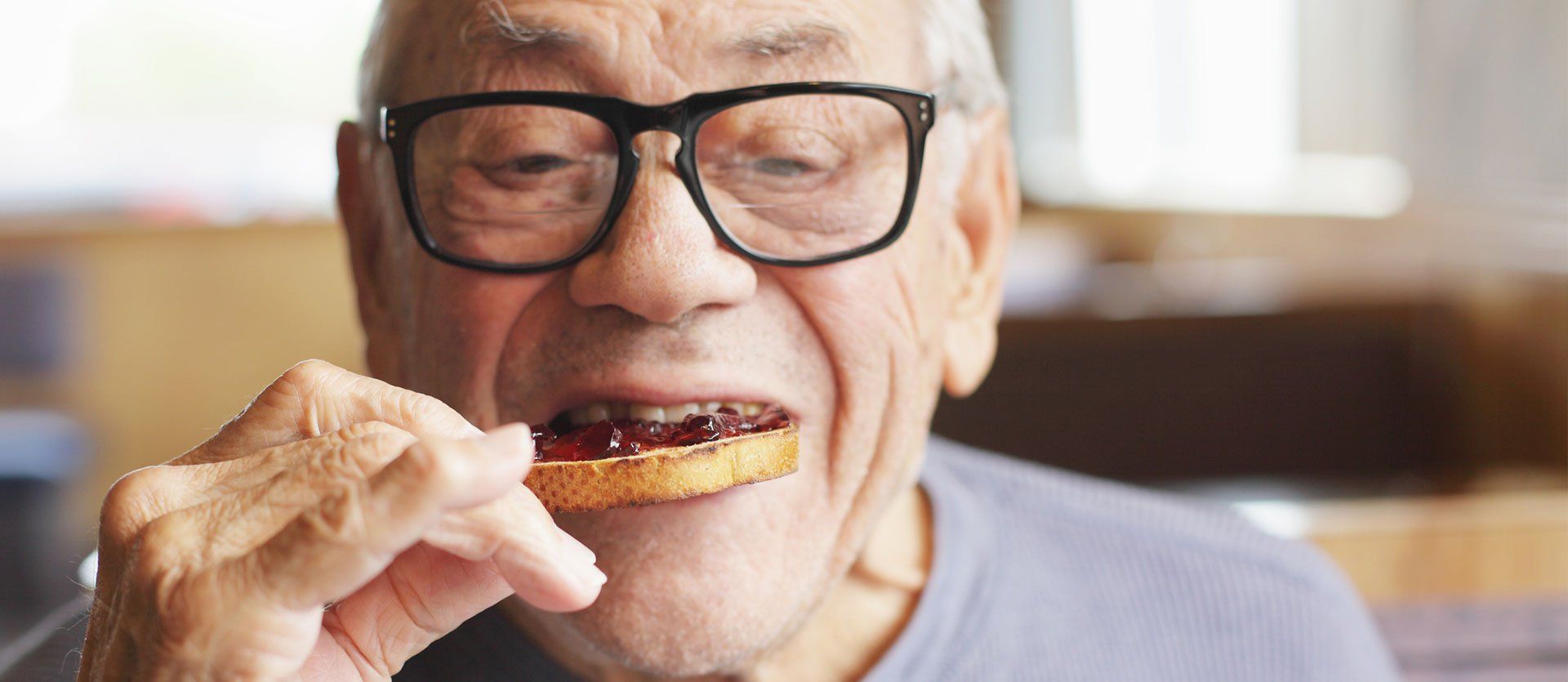 An elderly man wearing glasses is eating a piece of bread with jam on it.