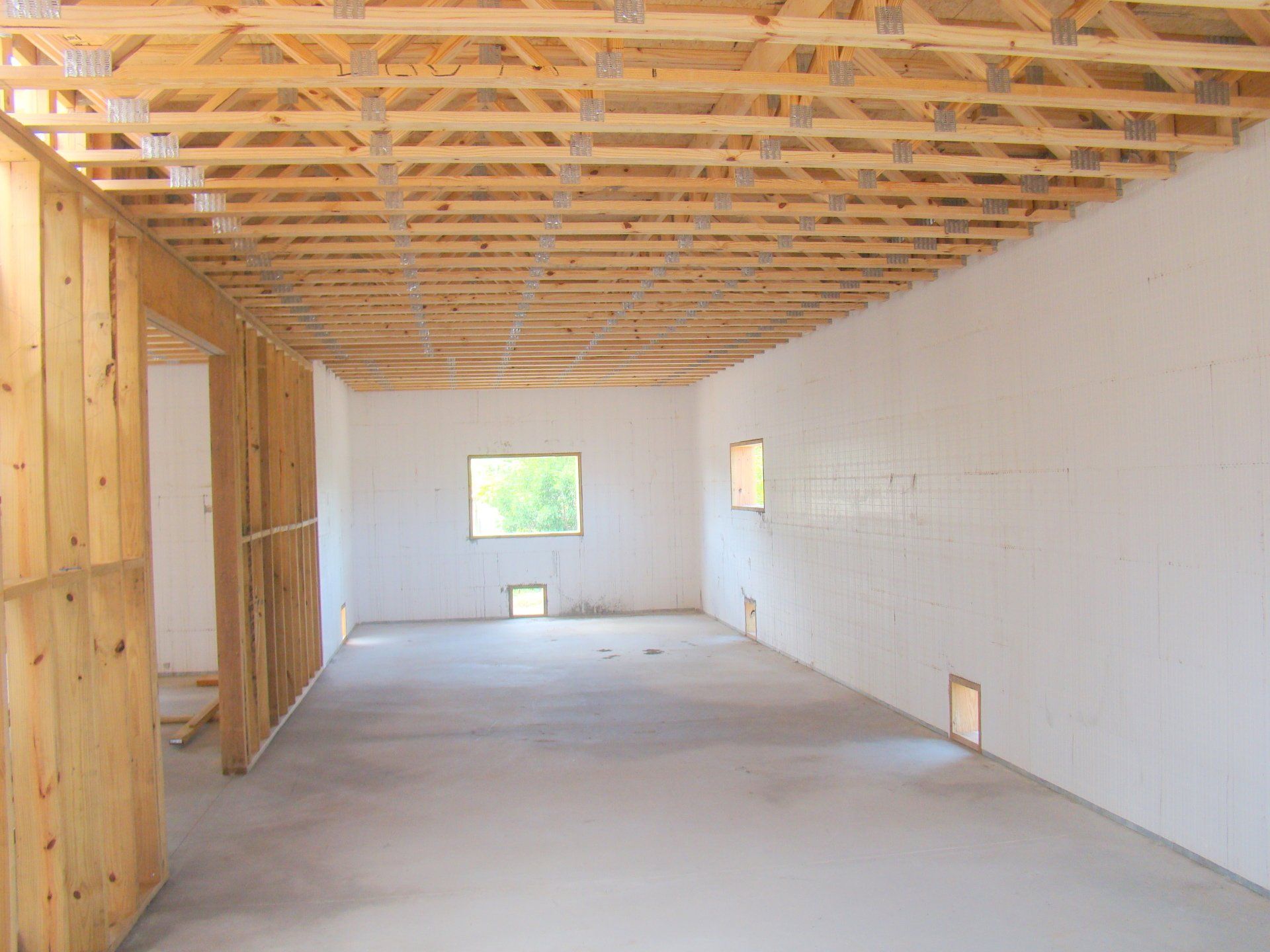 Interior of a room under construction with wooden framing, white walls, concrete floor, and windows.
