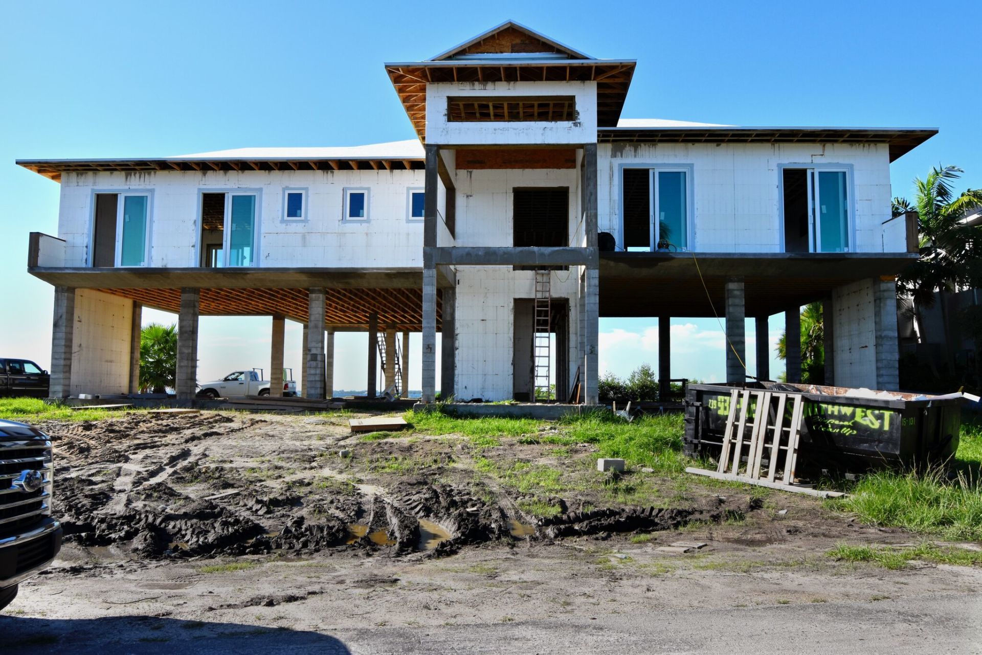 House under construction elevated on pillars; blue sky, mud and grass in front.