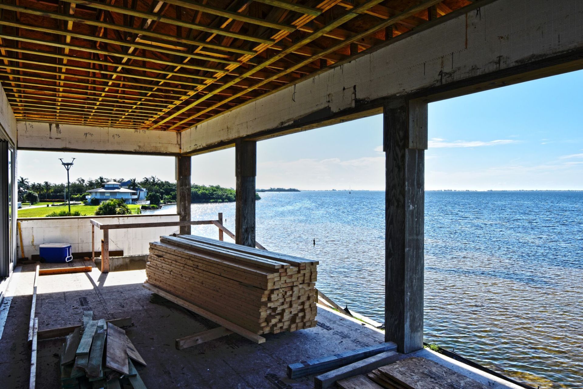 Construction site with view of water, stack of lumber, wood ceiling framing, concrete pillars, blue sky.
