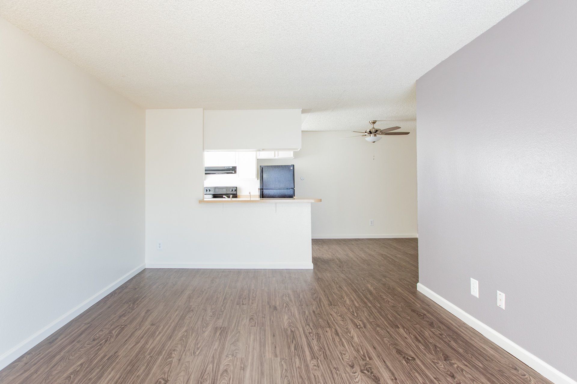 Empty modern living room with wood flooring, white walls, and a small pass-through kitchen counter.