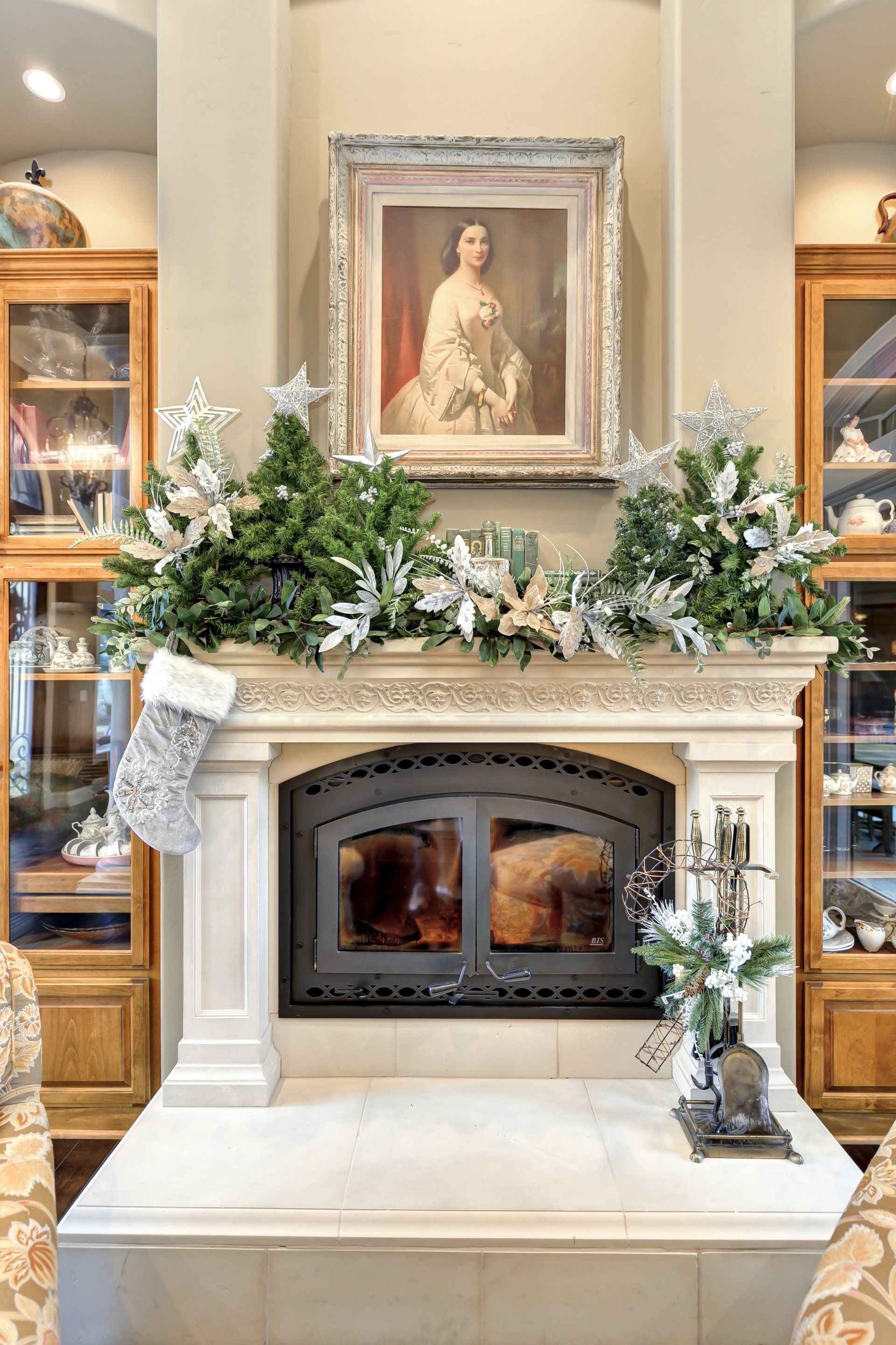 Fireplace decorated for Christmas with portrait above, flanked by glass-door cabinets.