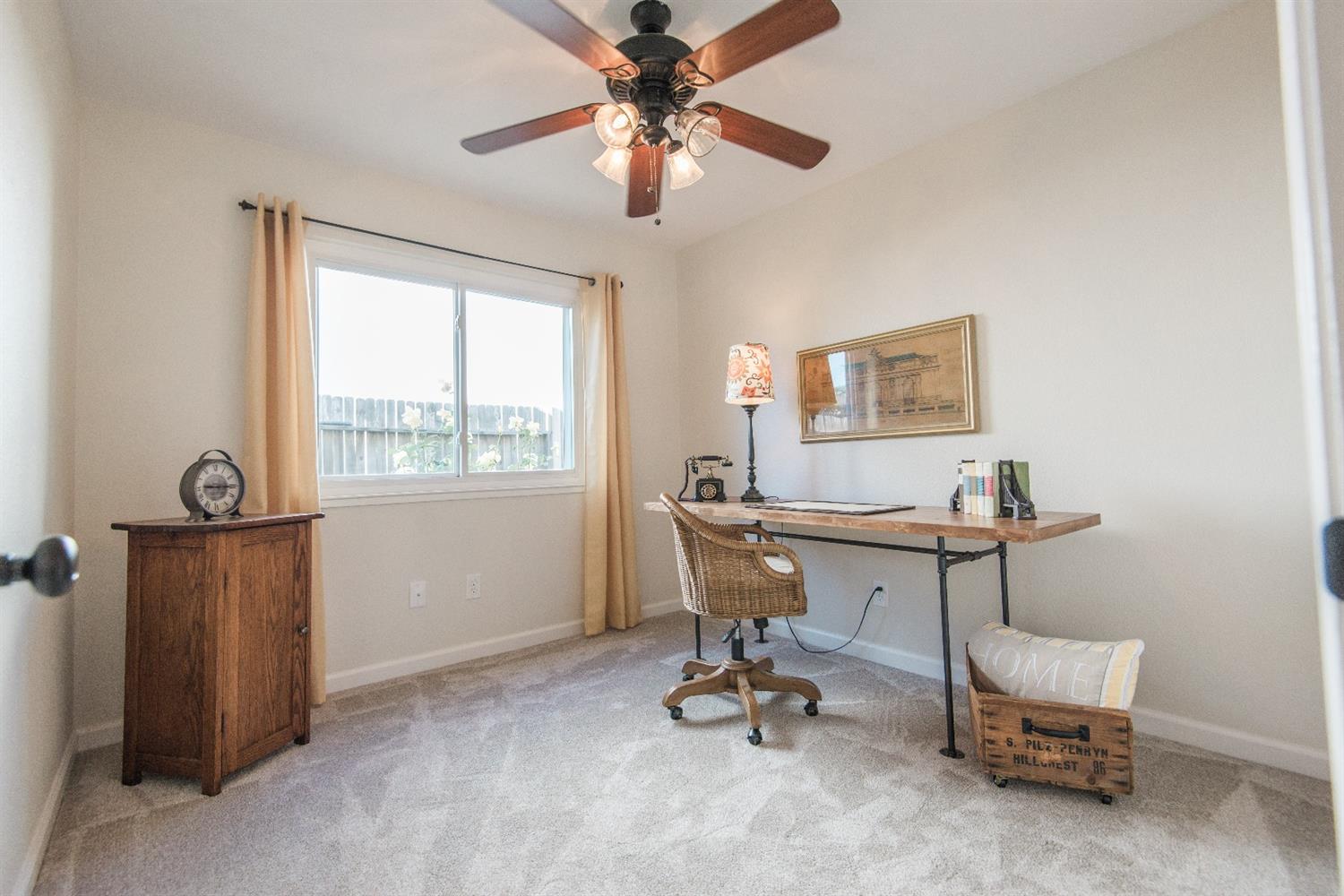 A home office with a desk, chair, and cabinet. The room has light walls, a window, and a ceiling fan.
