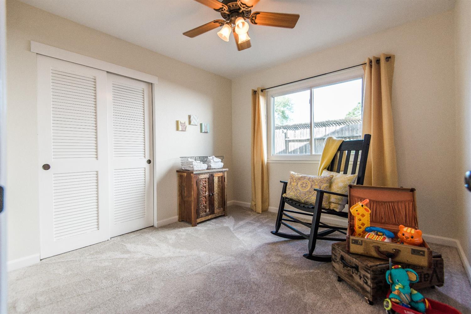 Cozy nursery with white closet doors, a rocking chair, and a small dresser. Neutral tones and a window with light filtering in.