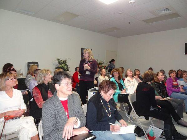 A woman speaks at a conference to a room of attentive women, some taking notes.