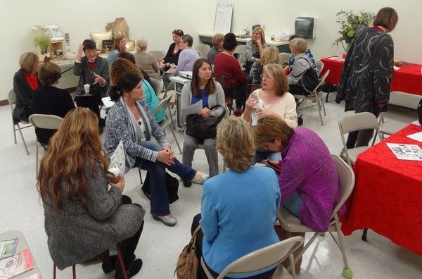 People seated in circles, conversing. Red tables and flowers in a bright room.