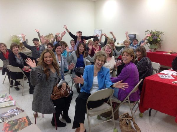 Group of women smiling and waving in a room, some sitting, others standing, all appearing happy.