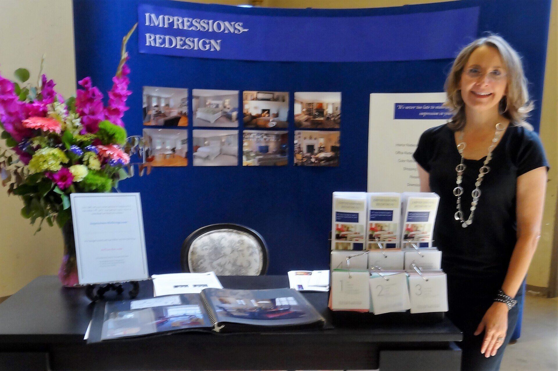 Woman at a trade show booth for home redesign, with photos, flowers, and products on display.