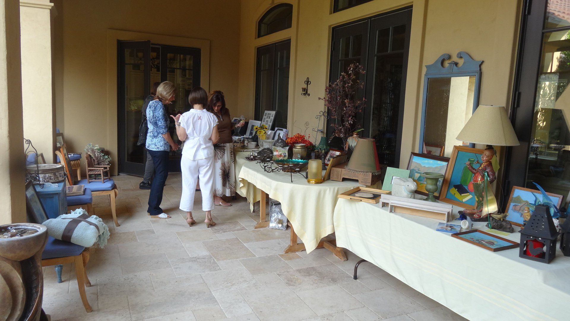 People browsing a table of items on an outdoor porch. Table has items for sale, beige walls, and stone floor.
