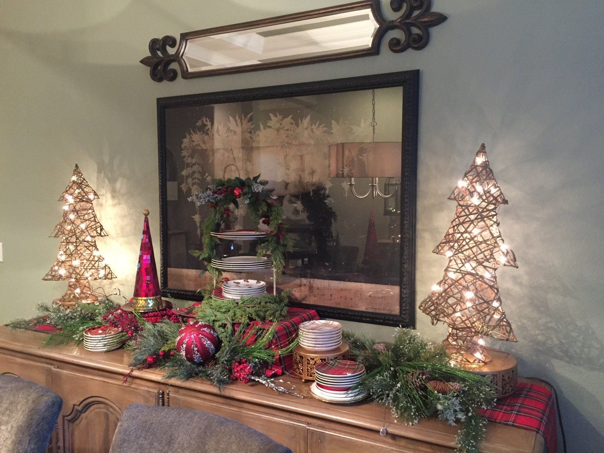 Christmas-decorated buffet: lighted trees, garland, ornaments, mirror, and greenery on a green wall.