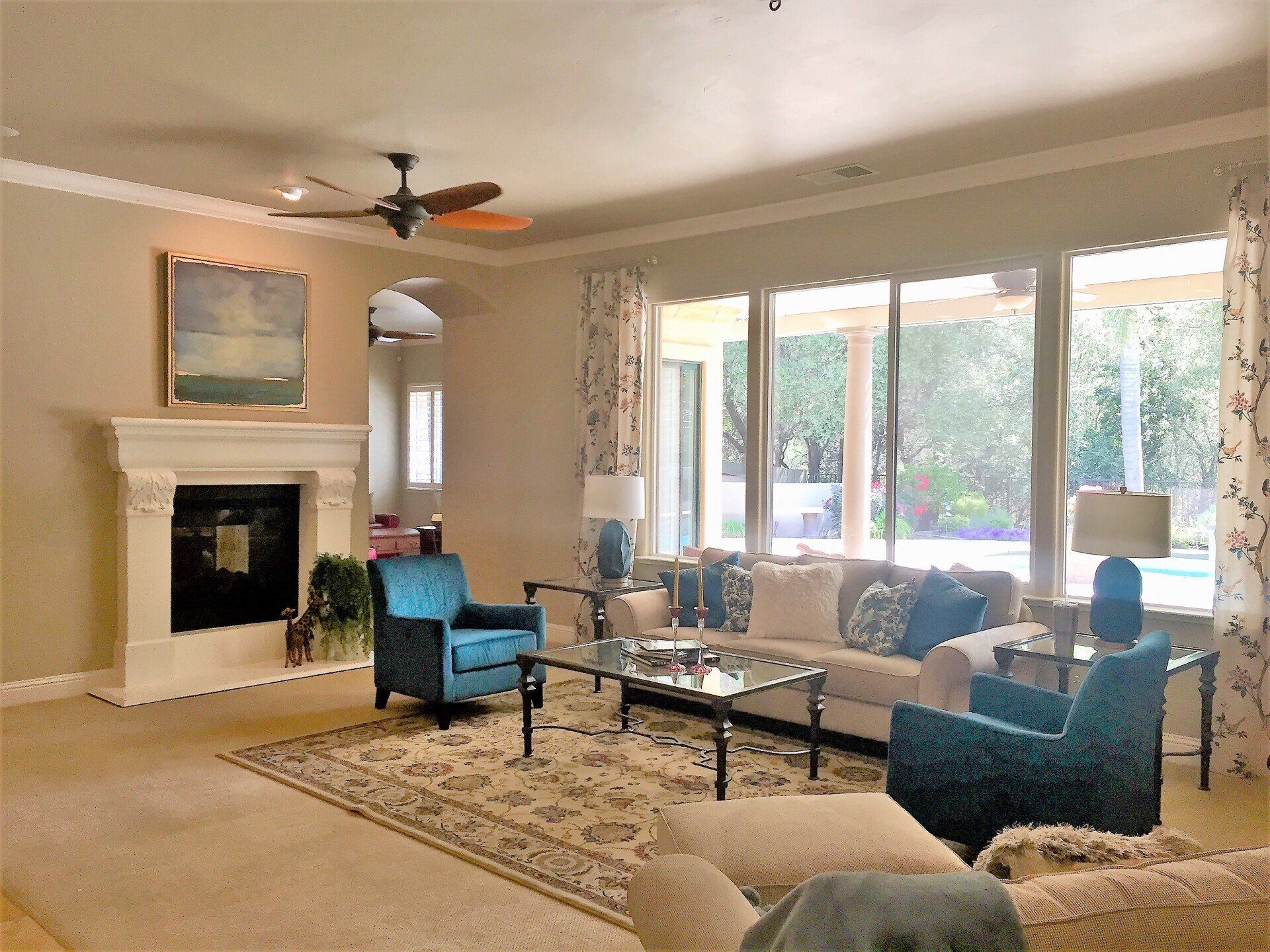 Living room with beige walls, fireplace, teal chairs, and large windows overlooking a yard.