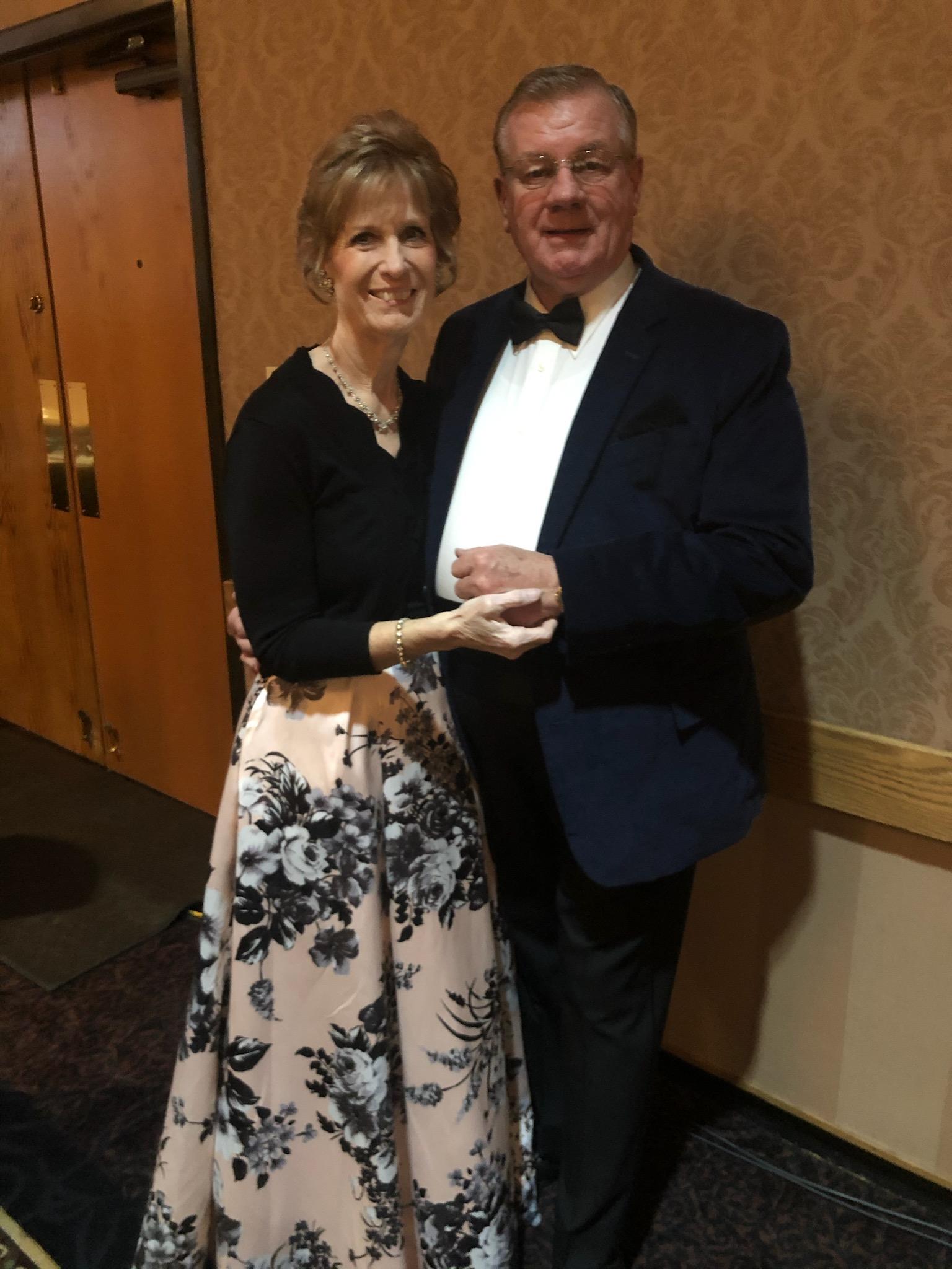 Couple in formal attire; woman in floral skirt, man in tuxedo, embracing, smiling indoors.