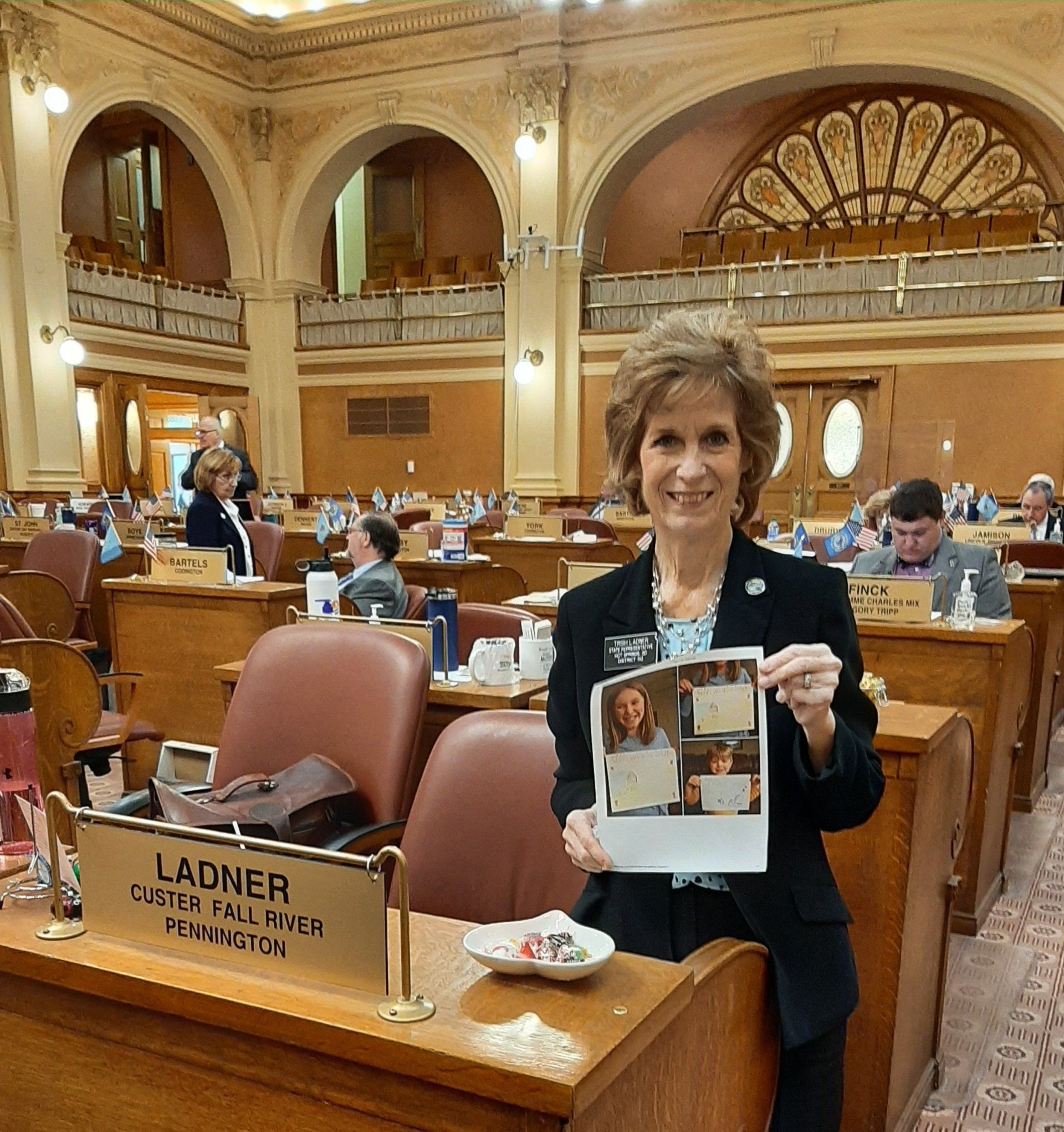 Woman holding printed photos in a legislative chamber, smiling. Nameplate reads 