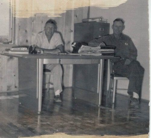 Two men seated at a desk, papers and binders present. Wooden paneled wall and shiny floor.