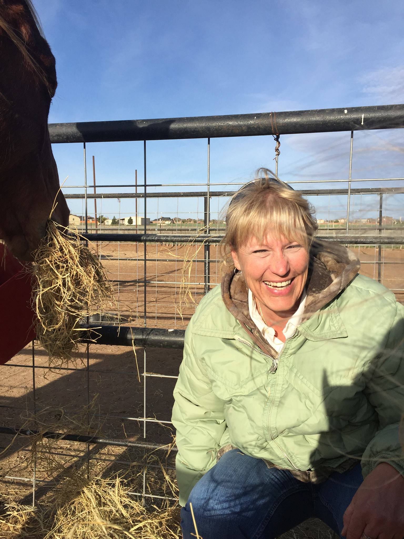 Smiling person in a light green jacket sits in a barn with hay and livestock fencing.