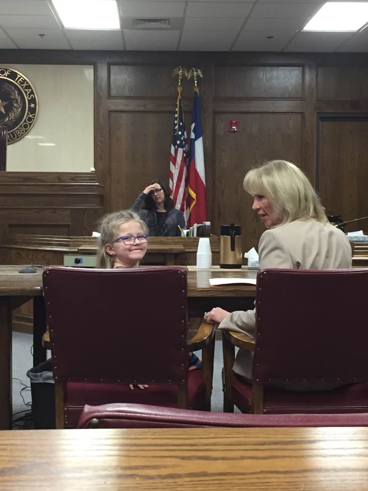 Two women seated in a courtroom, one turning to speak to the other; U.S. flag and judge’s bench in back.