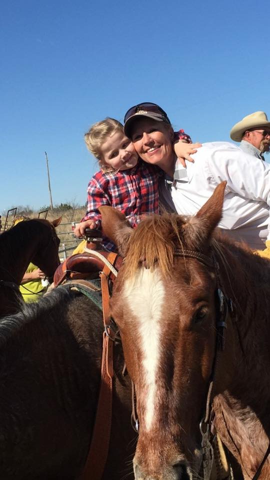 Child hugging an adult on a horse at an outdoor rodeo, with clear blue sky and spectators nearby