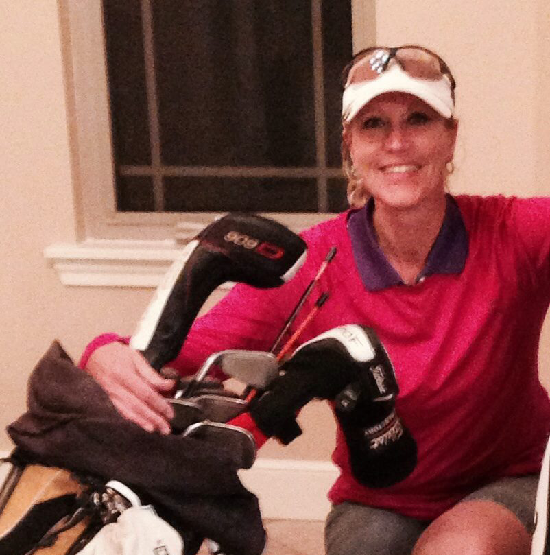 Woman smiling beside golf bag and clubs indoors, wearing a pink shirt and white visor.