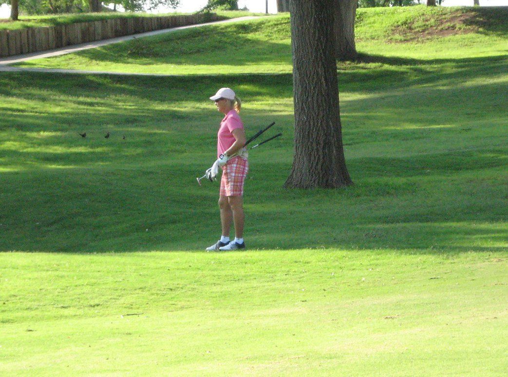 Golfer in pink stands on a green fairway beside a tree, holding a club.