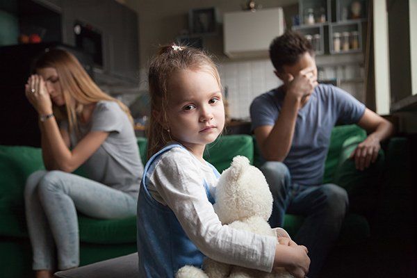 Child holding a teddy bear in the foreground, with two upset adults sitting on a couch in the background.