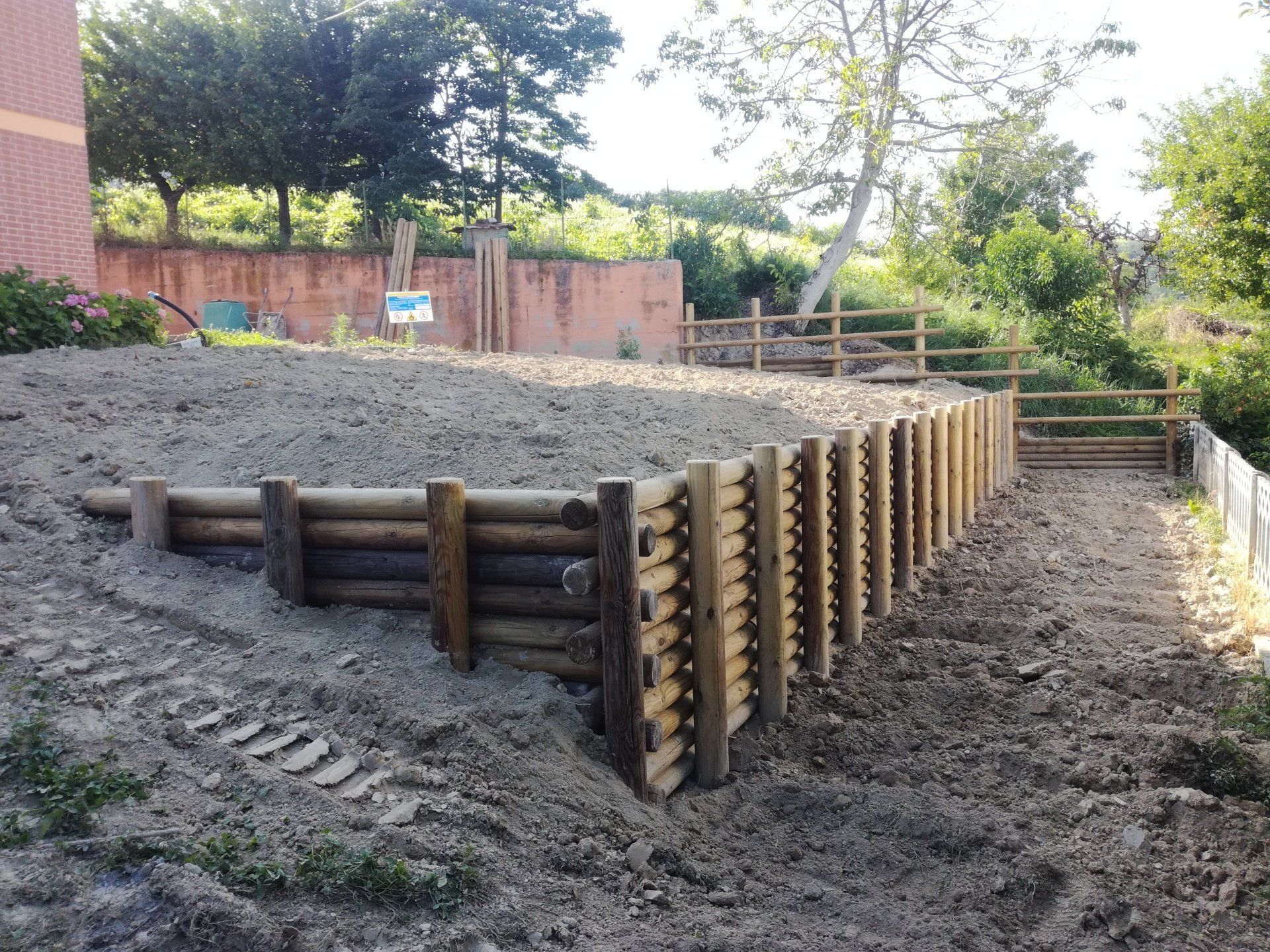 Muro di contenimento in legno su un pendio, con terra e recinzione in un ambiente esterno soleggiato.