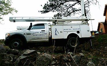 White Living Water Pump Service truck on a rocky hillside with its extendable arm extended, near a house.