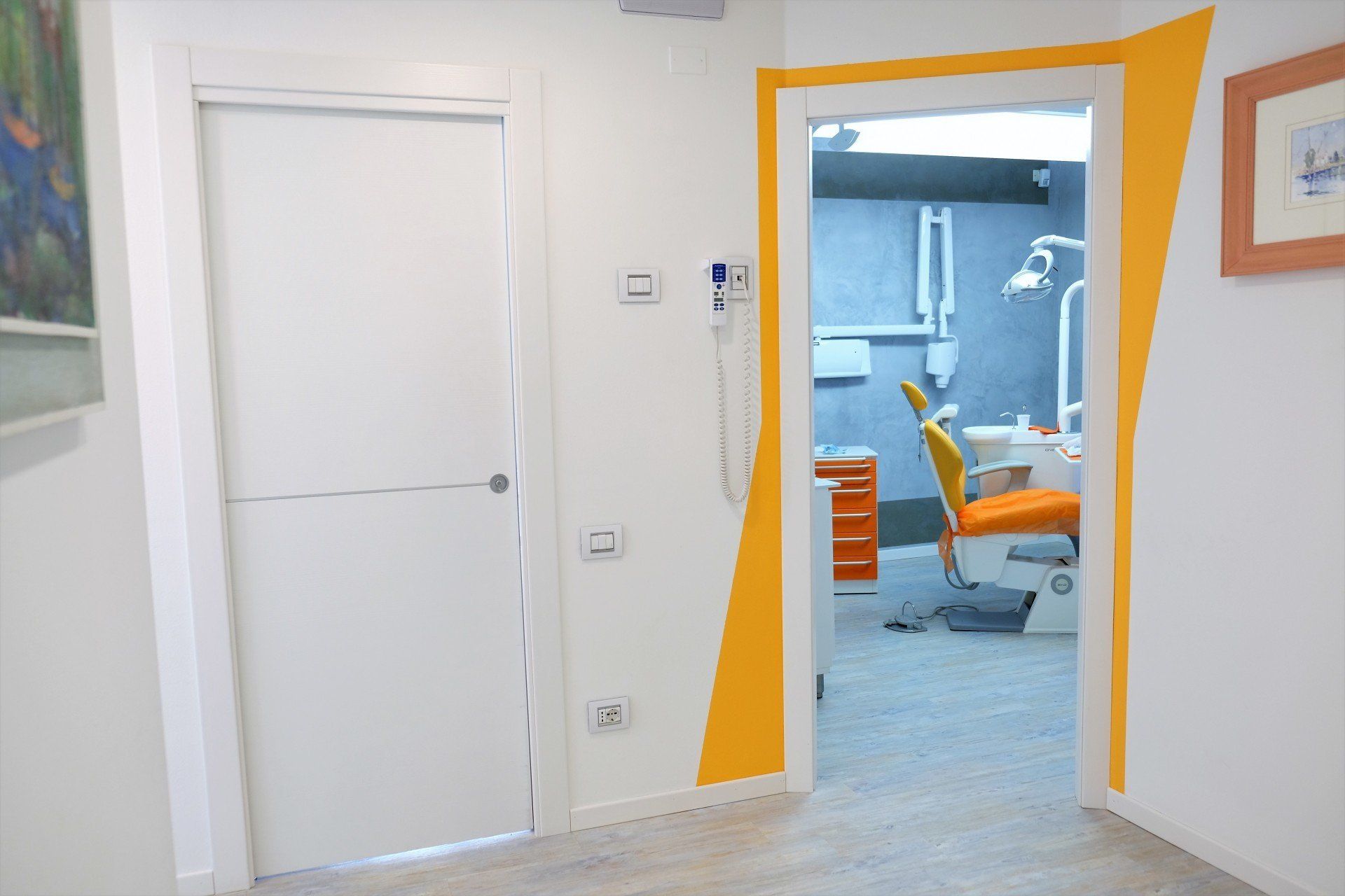 Corridoio con porta bianca a sinistra e porta d'ingresso con bordi arancioni che conduce allo studio dentistico a destra. Poltrona odontoiatrica visibile.