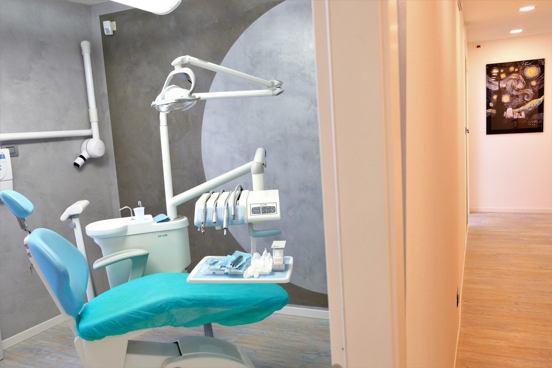 Sala visita dello studio dentistico con poltrona blu, attrezzatura, parete grigia e corridoio visibili.